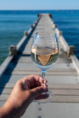Hand with glass of cold rose wine from Provence and wooden yacht boota pier on white sandy beach Plage de Pampelonne near Saint-Tropez, summer vacation in France