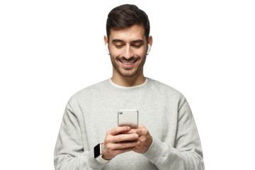 Young man standing, wearing wireless earphones, looking at screen of phone with smile, typing or surfing online