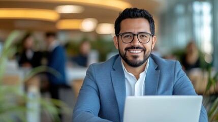A man in a blue blazer and glasses is smiling while sitting at a desk with a laptop in a modern office environment with blurred colleagues in background.