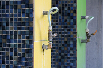 Close up of Two water faucets with hoses connected to them are mounted on a wall with blue ceramic tile and yellow and green paint. A close up of two outdoor faucets with blue and green tile walls