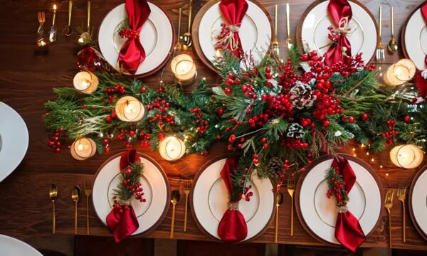 A beautifully set holiday dinner table adorned with red napkins, evergreen centerpieces, candles, and festive decorations, creating a warm and inviting Christmas atmosphere