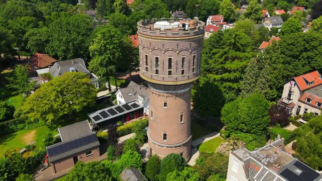 Experience an aerial perspective of Baarn's historic watertower, surrounded by lush greenery and cozy residential homes, showcasing the unique charm of this Dutch town.