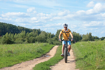 Cyclist in helmet with equipment on electric bicycle rides along path in field in countryside. Sports and health. Summer hobby. 