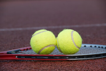 Closeup of a red tennis racket lying on a clay court with two tennis balls