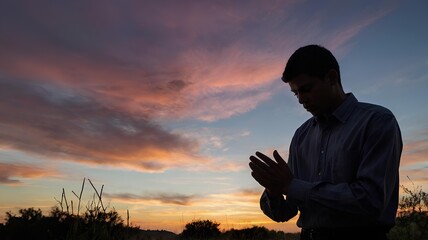 Fototapeta premium A young man’s silhouette on his knees, praying with clasped hands, set against a vibrant sunset, symbolizing sacrifice and faith.