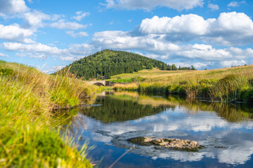 Bukovec Mountain mirrors on a tranquil stream surrounded by grassy fields and scattered clouds in Jizerka Village, Jizera Mountains of Czechia.