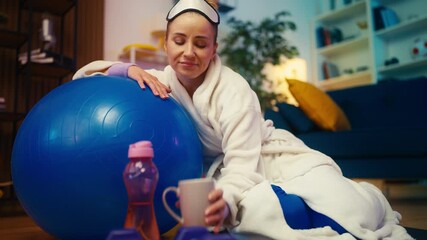 Woman in bathrobe leaning on fitness ball and drinking coffee, missed workout