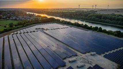 Aerial view of a large solar farm with wind turbines in the background, set against a scenic...