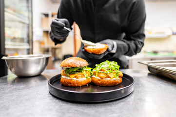 A chef in a black uniform assembles a gourmet burger with fresh ingredients on a black plate in a professional kitchen. The close-up highlights culinary skills and attention to detail.