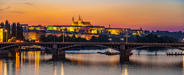 Fototapeta premium Jirasek bridge crosses the Vltava River in Prague, with Prague Castle illuminated on the opposite bank. The setting sun casts a warm glow on the cityscape, creating a serene and picturesque scene.