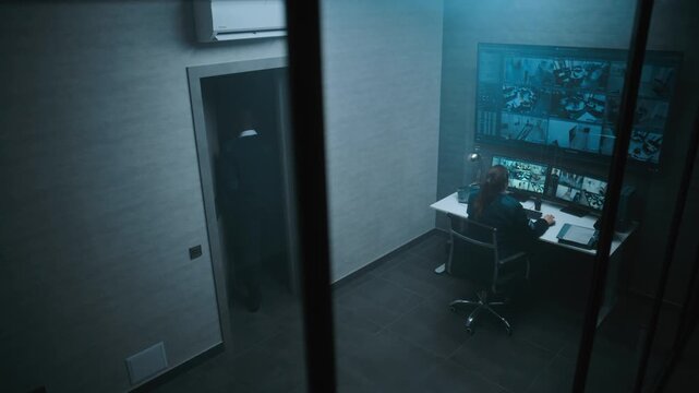 Female CCTV operator controls security cameras in monitoring center on computer. Surveillance video footage displayed on multiple monitors and big digital screen. African American coworker passes by.