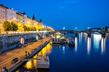 Fototapeta premium The Vltava River in Prague is bustling with activity at night. People are gathered on the embankment, enjoying the cool evening air. The city lights reflect off the water, creating a beautiful and
