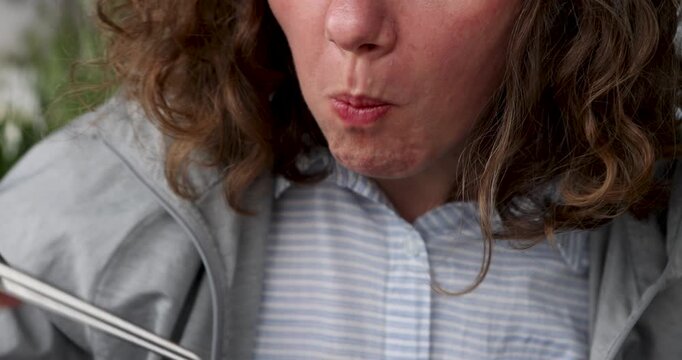 Close-up of curly woman eating pho bo soup with chopsticks using her mouth. Closeup image showcases a woman with curly hair in a light gray jacket over a striped shirt.