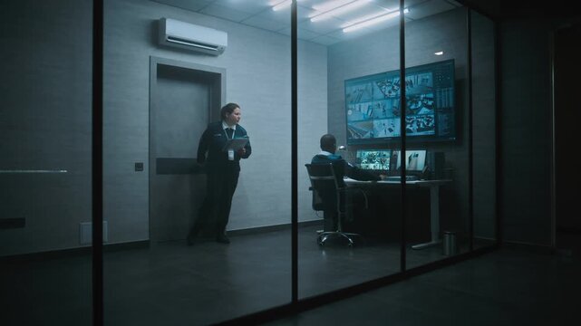 African American security operator monitors CCTV cameras in office on computer. Multiple monitors and big digital screen showing surveillance video footage. Female worker comes and talks to colleague.