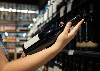Close-up of a hand holding a wine bottle in a store aisle with shelves of wine bottles in the background