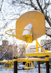 Yellow basketball hoop with a torn net is covered in snow on an empty playground, highlighting the stillness of outdoor spaces during winter