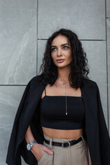 Pretty fashionable young beauty woman with curly hair in trendy top with black blazer stands near a gray building on the street