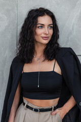 Fashion happy young beauty curly woman with black blazer in top stands near a gray background outdoors