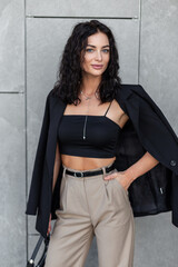 Fashionable beautiful woman with curly hair in black blazer and top with pants stands near a gray wall on the street. Urban pretty lady