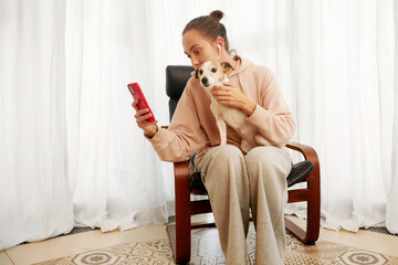 Young woman sits in a modern chair, holding her disabled dog and using her smartphone. The bright room with white curtains and a stylish rug creates a cozy, inviting atmosphere for her peaceful moment