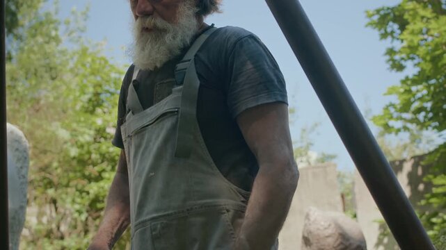Medium shot of mature Caucasian male sculptor pulling chains while working with heavy natural stone outdoors in garden