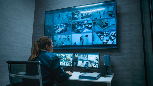 Female CCTV operator controls office surveillance cameras with face recognition using computer, works in modern monitoring center. Multiple monitors and big digital screen show security video footage. - Powered by Adobe