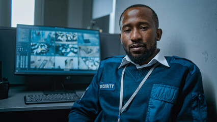 African American male security guard sits at the workplace in the office and looks at camera. Computer monitor with displayed surveillance video footage from CCTV cameras in the background. Portrait.