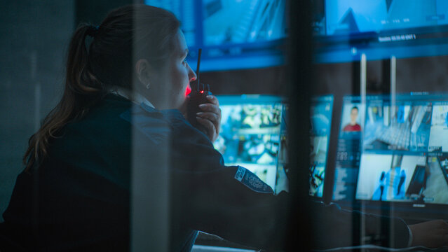 Close up of female CCTV operator working on computer in monitoring center with security cameras, talking on walkie talkie. Multiple monitors and big digital screen showing surveillance video footage.