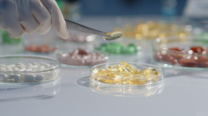 Many petri dishes containing different types of pills on a laboratory table. The researcher's hand is picking up a transparent fish oil capsule. Scenes for advertising medicine and functional foods