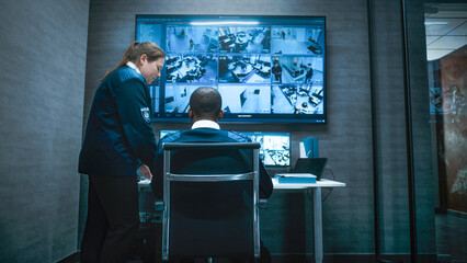 Back view of African American security worker controlling CCTV cameras on computer. Multiple monitors and big digital screen show surveillance footage. Female employee comes in and talks to operator.