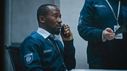 African American security officer talks in walkie talkie. Female coworker stands nearby and uses tablet computer. Multiethnic surveillance operators control CCTV cameras in modern monitoring center.