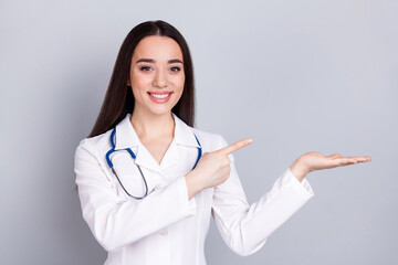 Portrait of qualified doctor woman point finger hold empty space white coat isolated on grey color...