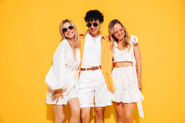 Fototapeta premium Group of young three stylish friends posing in studio. Fashion man and two cute female dressed in casual summer clothes. Smiling models having fun. Cheerful women and guy near yellow wall, sunglasses