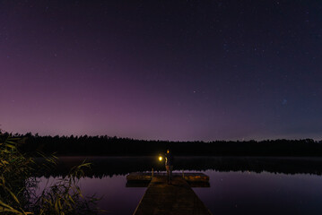 Woman with a lantern on a wooden pier, Aurora Borealis northern lights, Jupiter, Mars, stars over the forest lake in Latvia on August night. © Julija