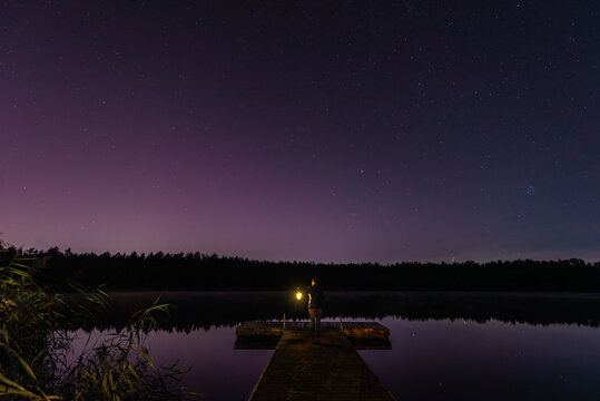 Woman with a lantern on a wooden pier, Aurora Borealis northern lights, Jupiter, Mars, stars over the forest lake in Latvia on August night.