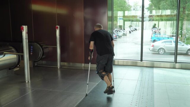 Mature man with bondage, medical splint, knee brace on the leg goes down the escalator to the subway using crutches. Accessible urban environment for the disabled, recovery from injury.
