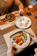 handsome girll is eating delicious, juicy baked fish with fresh vegetables, fragrant herbs, and avocado butter sauce, served on a beautiful white plate on a wooden table in a cozy, charming restaurant