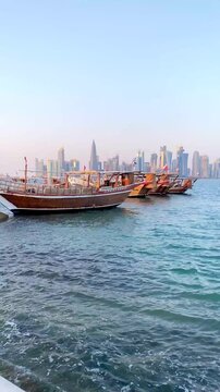 Traditional Arab dhow boats are anchored on the water near the shore of the bay, against the backdrop of the skyline of Doha.