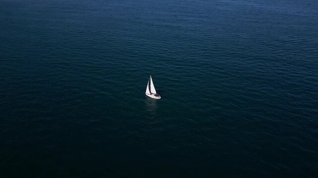 A serene sailing boat glides on calm, deep blue waters under a clear sky. Distant ships and sailboats dot the horizon, creating a peaceful maritime scene