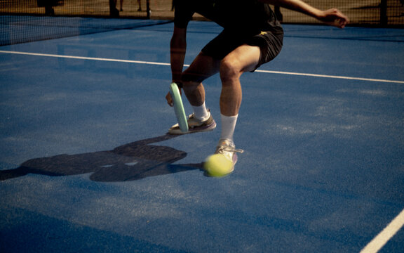 Male padel player in action on blue tennis court with light blue racket, playing in full sun with harsh shadows