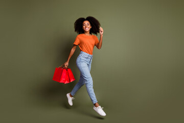 Full length photo of charming positive woman wear orange t-shirt walkign rising shoppers empty space isolated khaki color background