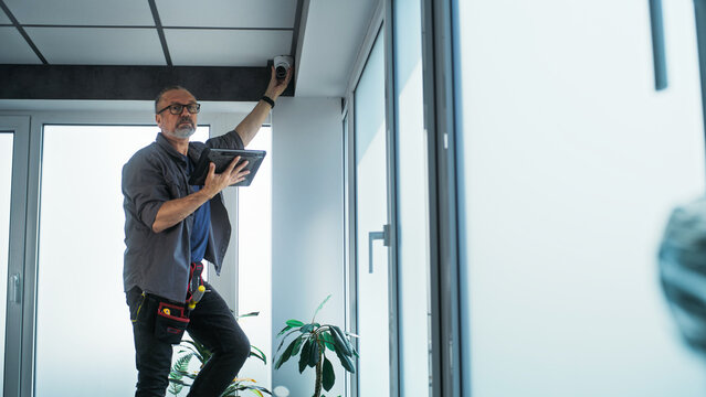 Male mature technician stands on ladder, installs CCTV camera in office room using tablet computer with professional software. Monitoring and tracking. Security system, video surveillance and privacy.