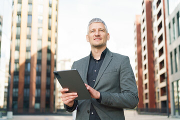 A businessman in a gray suit confidently holds a tablet, looking ahead in a modern urban environment.