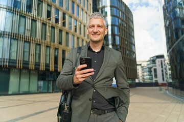 A businessman in a gray suit smiles while checking his phone, carrying a bag in a modern urban environment.