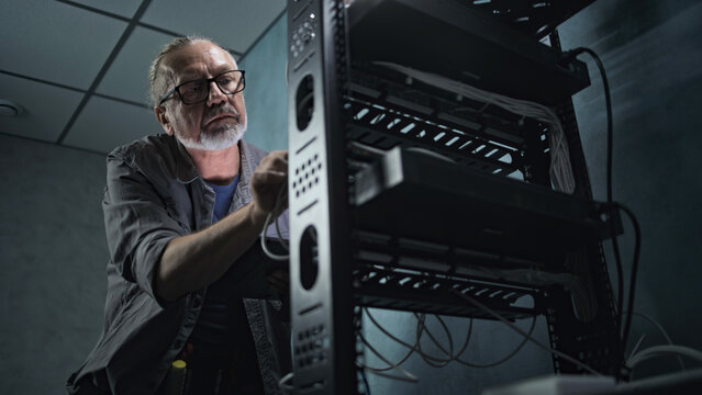 Mature engineer inserts the cable into the communications equipment, uses tablet computer. Male system administrator works in server room. Local area network or web server maintenance. Tech support.