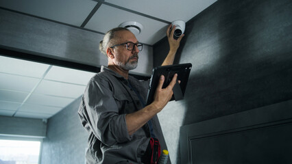 Male installer adjusts the angle of CCTV camera in office, sets up it using digital tablet computer...