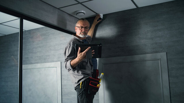 Male installer adjusts the angle of CCTV camera in office, sets up it using digital tablet computer with professional program. Monitoring and tracking. Security system, video surveillance and privacy.