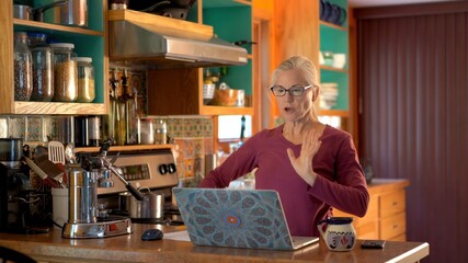 Mature woman talking to computer while working on computer in kitchen.