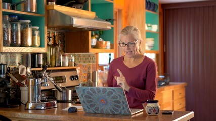 Mature woman talking to computer while working on computer in kitchen.
