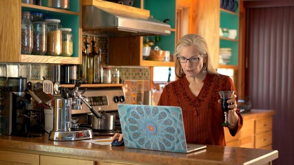 Shocked and concerned, a mature woman looks at her laptop while it rests on a kitchen counter.